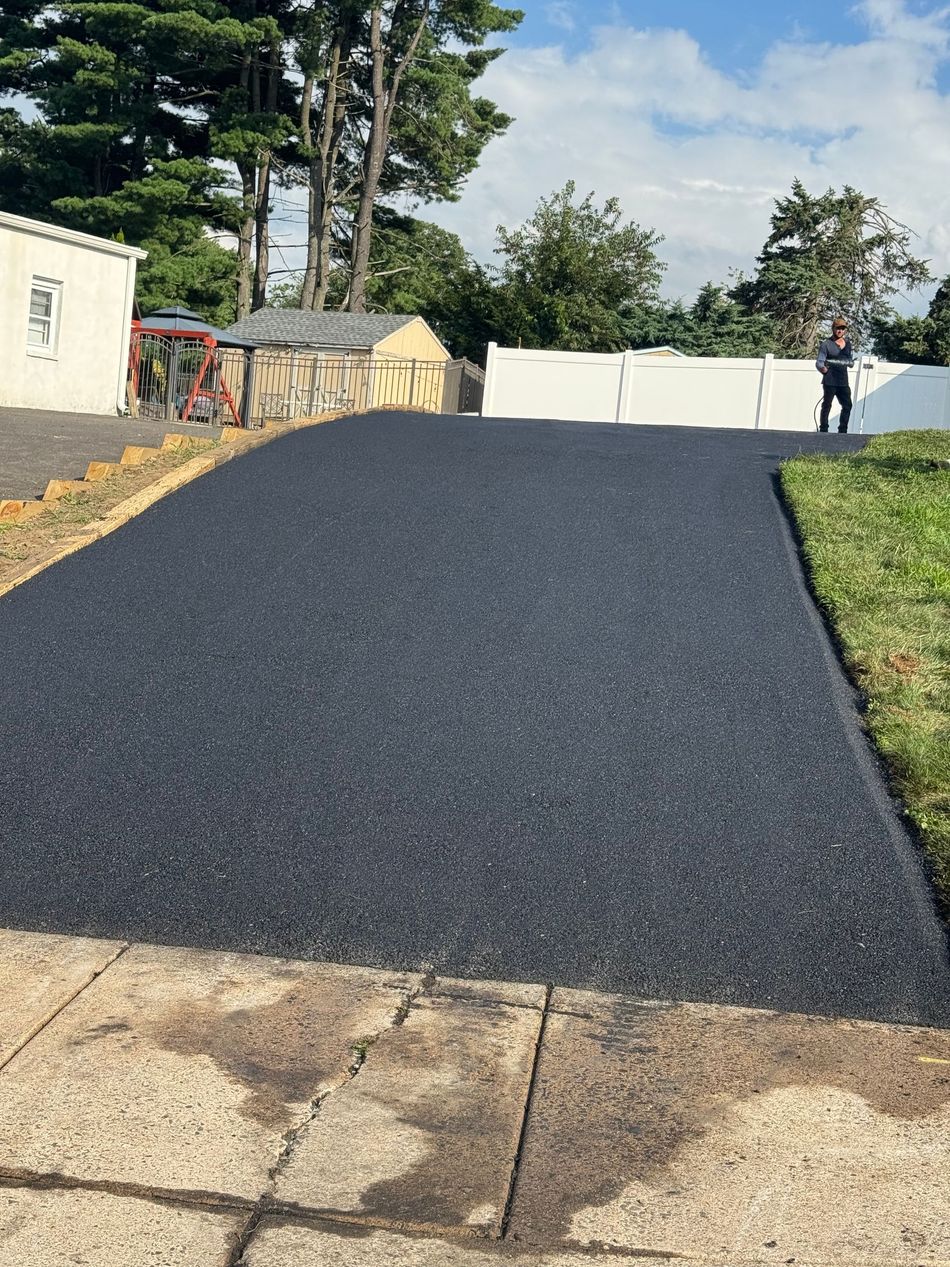 Freshly paved black driveway sloping up to a white gate, with a person standing in the distance.
