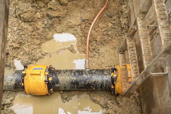 Yellow-and-black pipe coupling in muddy trench beside brick wall and ladder