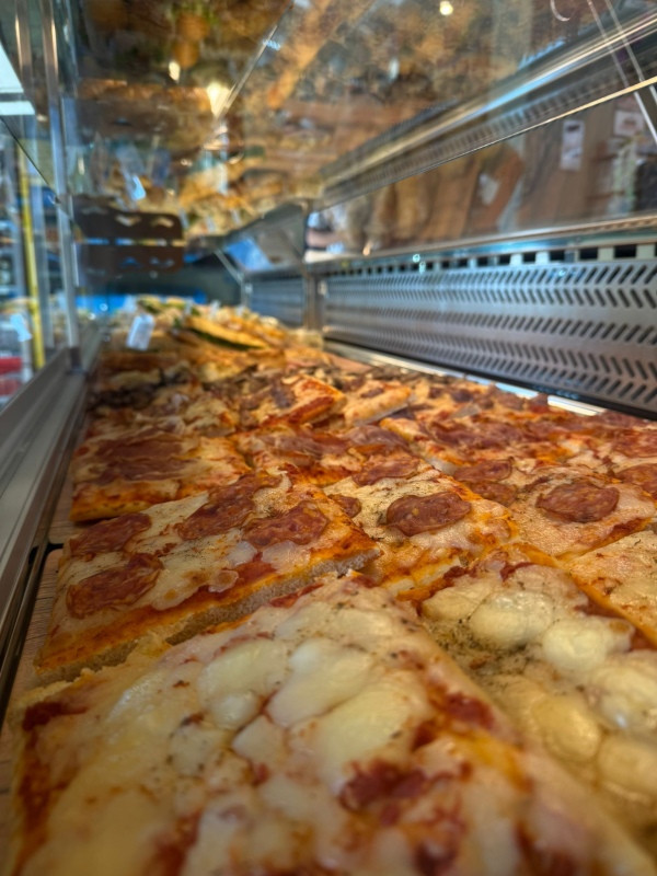 Primo piano di fette di pizza rettangolari in una vetrina; sono visibili il salame piccante e il formaggio.