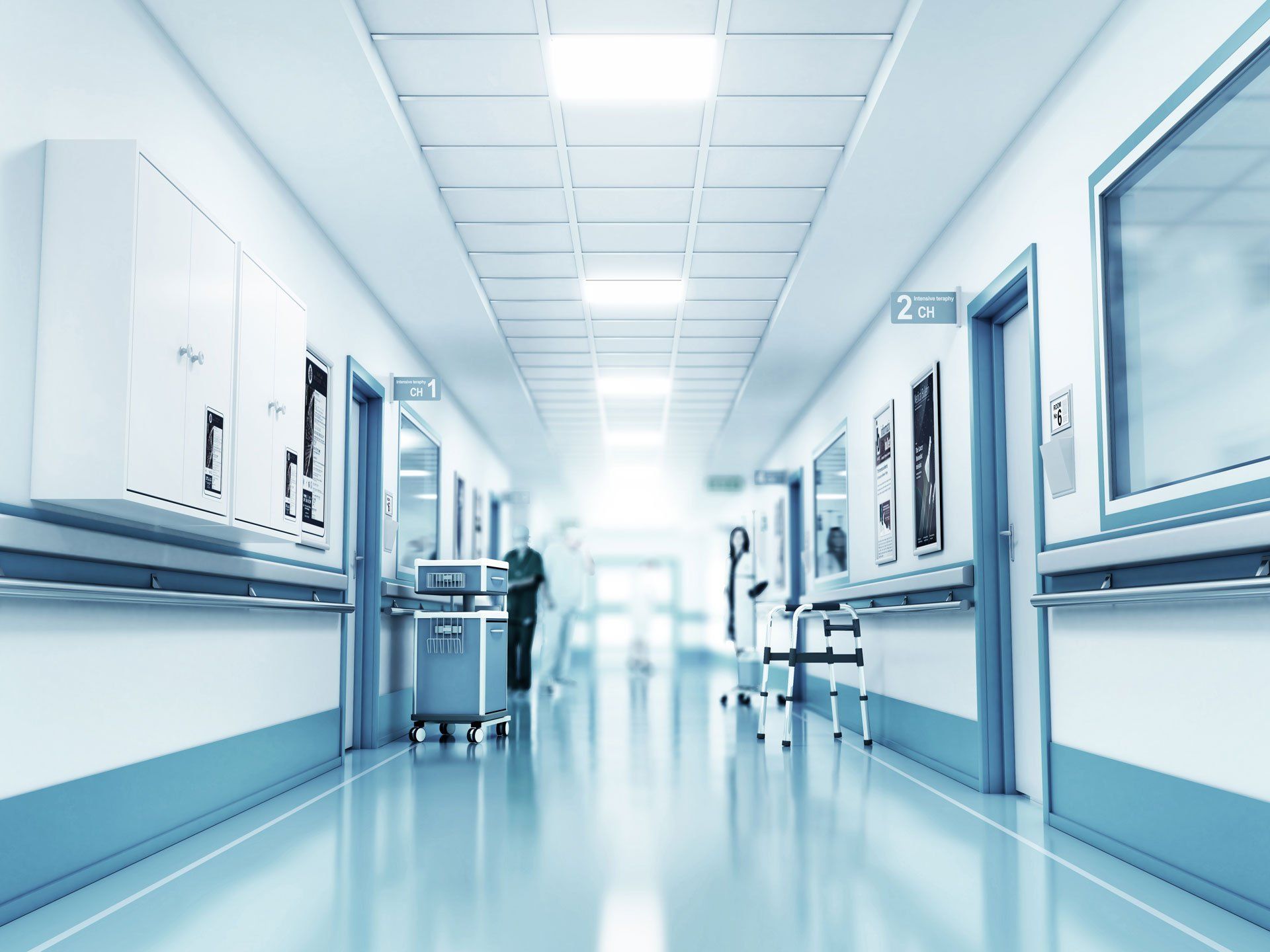 Bright, sterile hospital hallway with white walls, blue trim, medical equipment, and blurred figures in the distance.