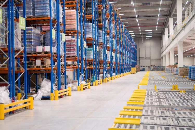 Tall blue storage racks filled with goods in a large warehouse with yellow safety barriers on a polished concrete floor.