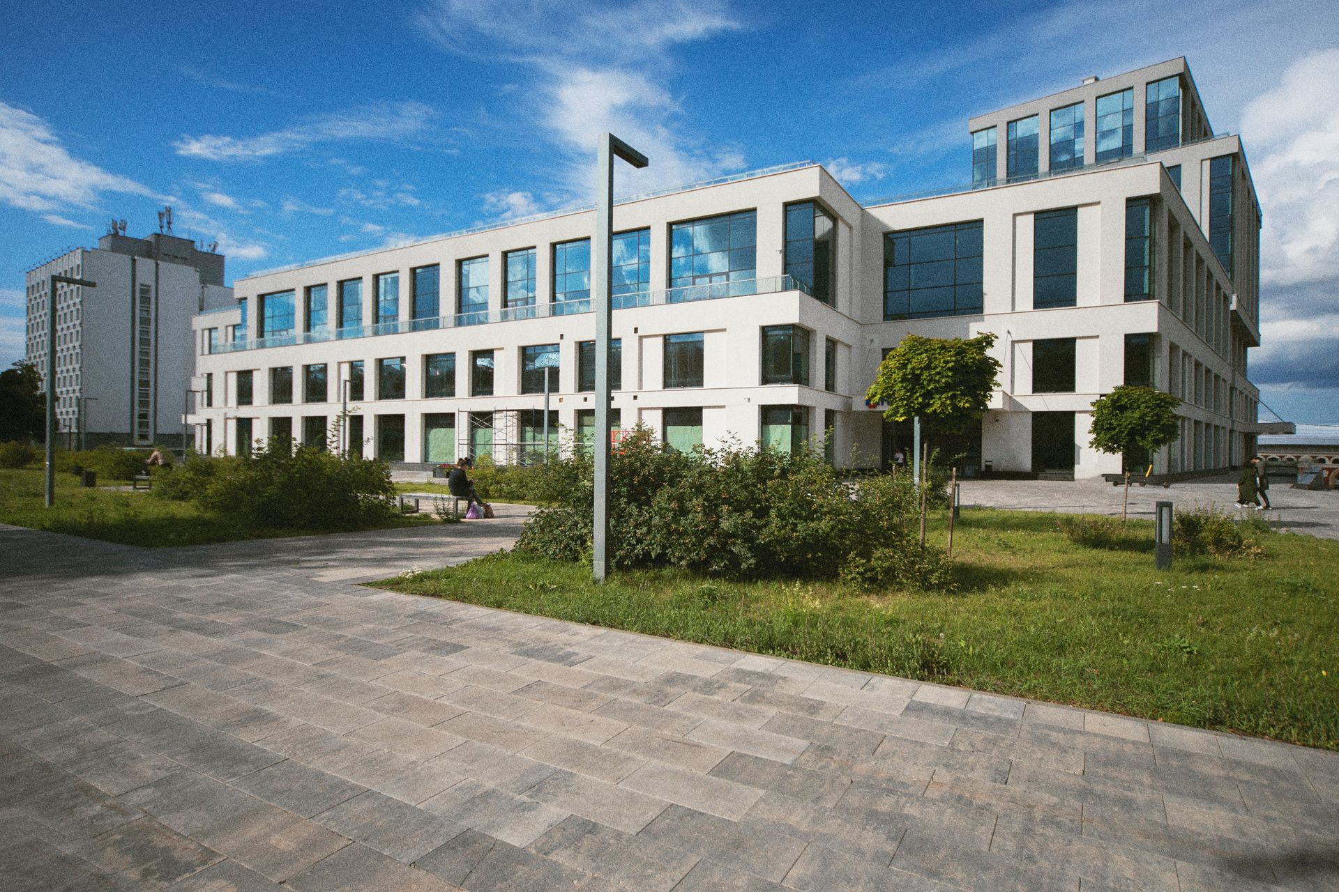 A white, modern office building with glass windows, set against a blue sky with a stone walkway and lawn in the foreground.