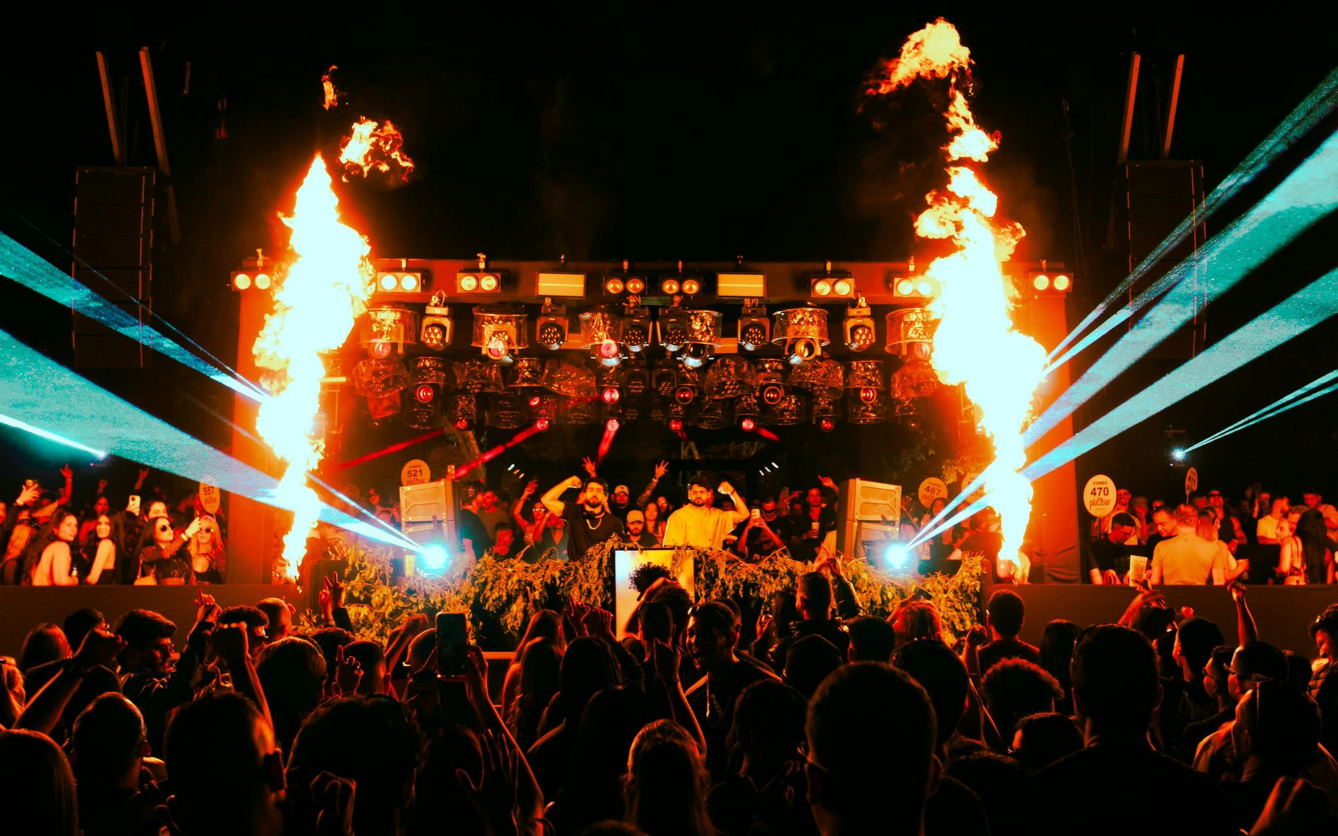 Concert with DJ on stage, pyrotechnics, and a crowd. Flames erupt, teal lights shine, and people dance.