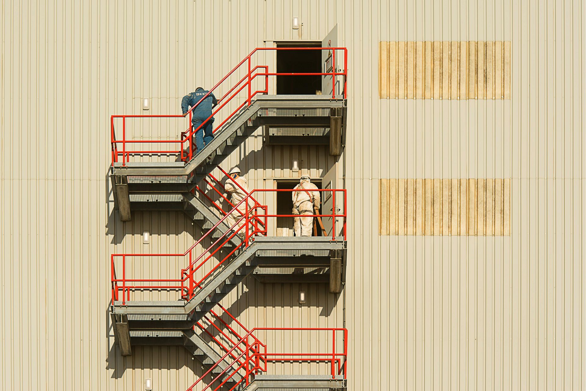 Workers in safety gear on a metal exterior staircase attached to a large, light-colored industrial wall. Workers in safety gear on a metal exterior staircase attached to a large, light-colored industrial wall.