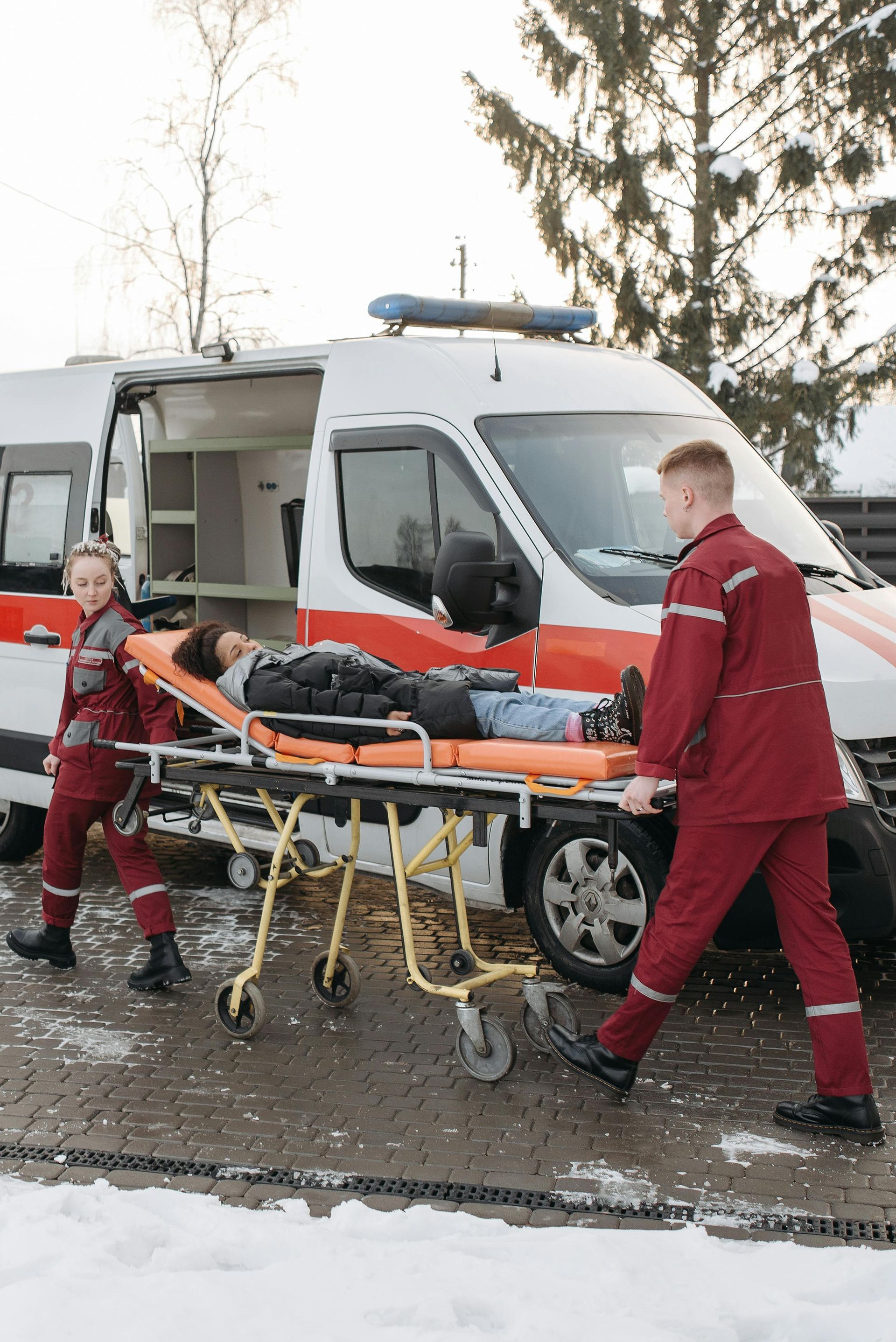 Two paramedics carrying patient on stretcher toward ambulance. Snowy outdoor setting.