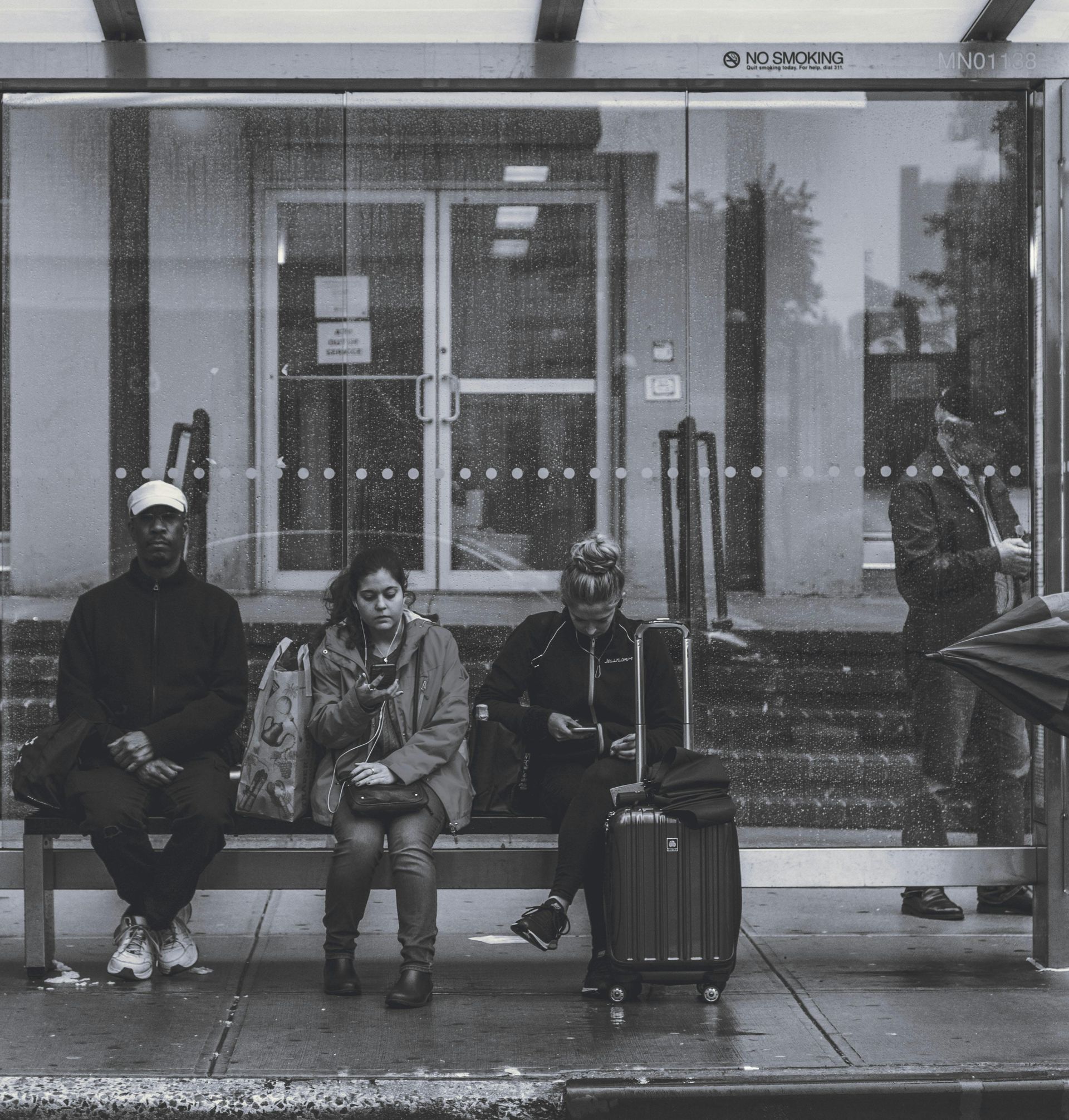 People waiting at a bus stop. Rain streaks the glass. A suitcase sits beside a person. One person looking at their for to see when the bus arrives with de Lijn app. People waiting at a bus stop. Rain streaks the glass. A suitcase sits beside a person. One person looking at their for to see when the bus arrives with de Lijn app.