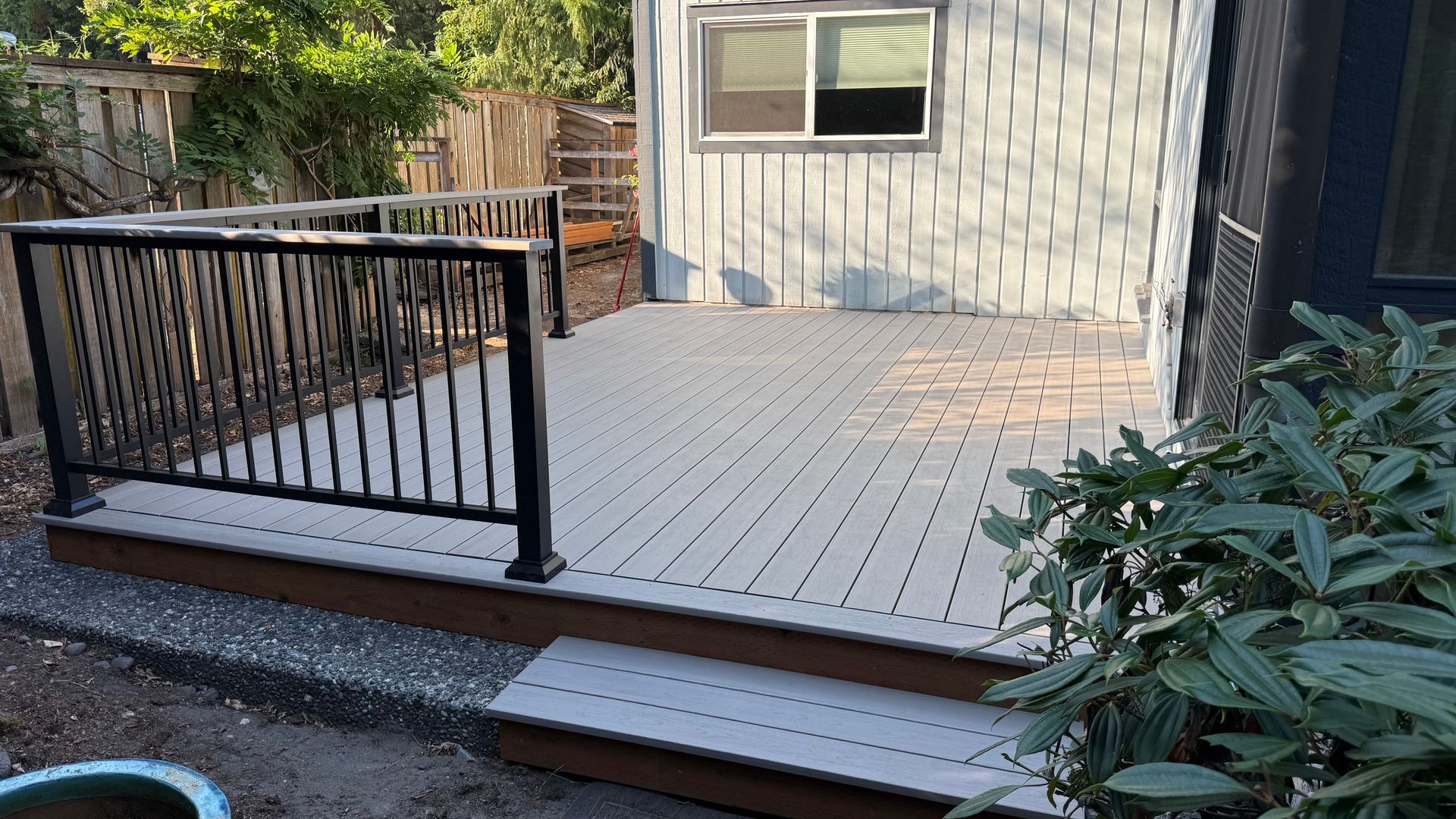 Small wooden deck with black railing beside a house, viewed from a garden path.
