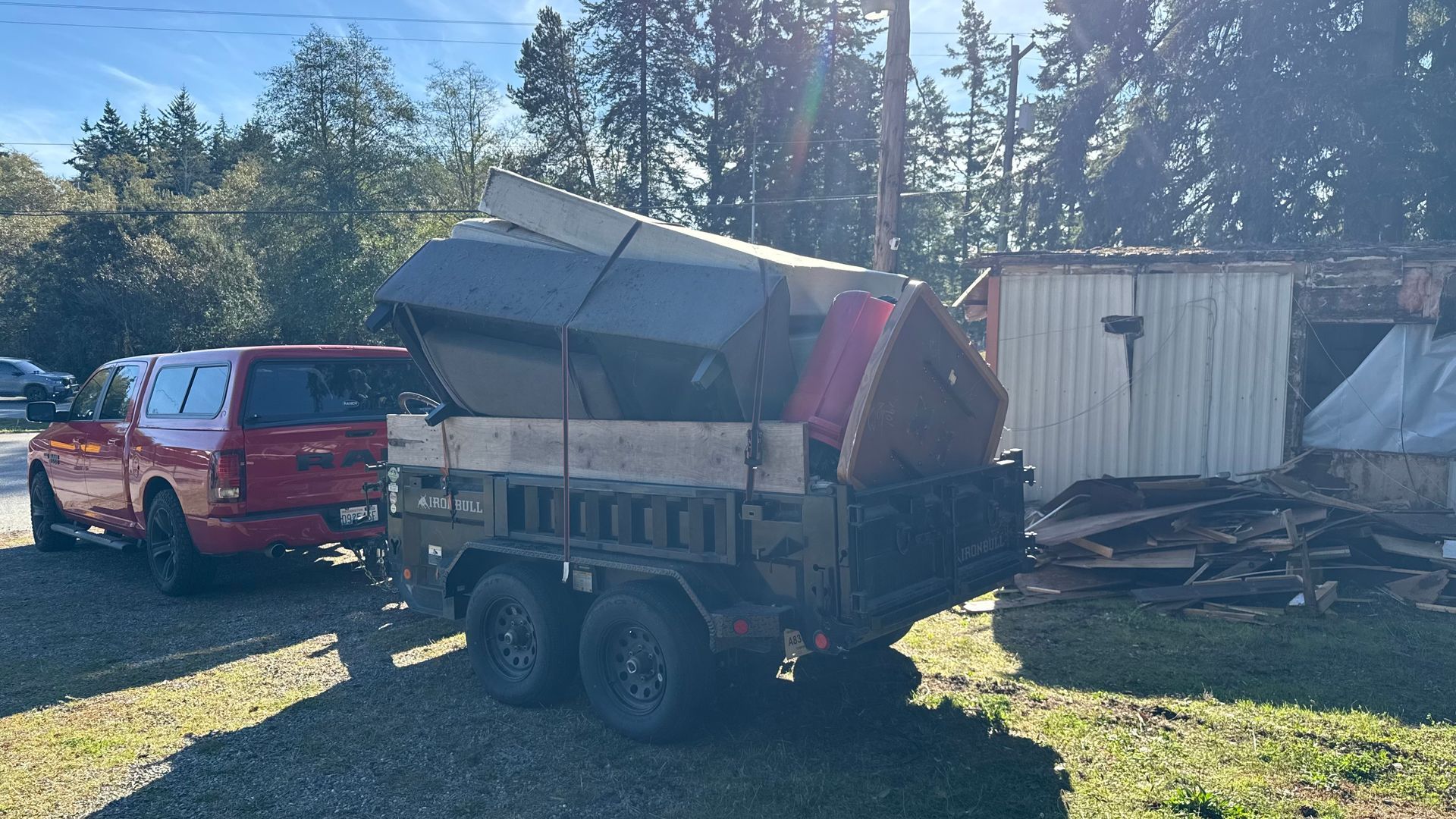 Red pickup and dump trailer loaded with debris beside a damaged shed in a grassy yard
