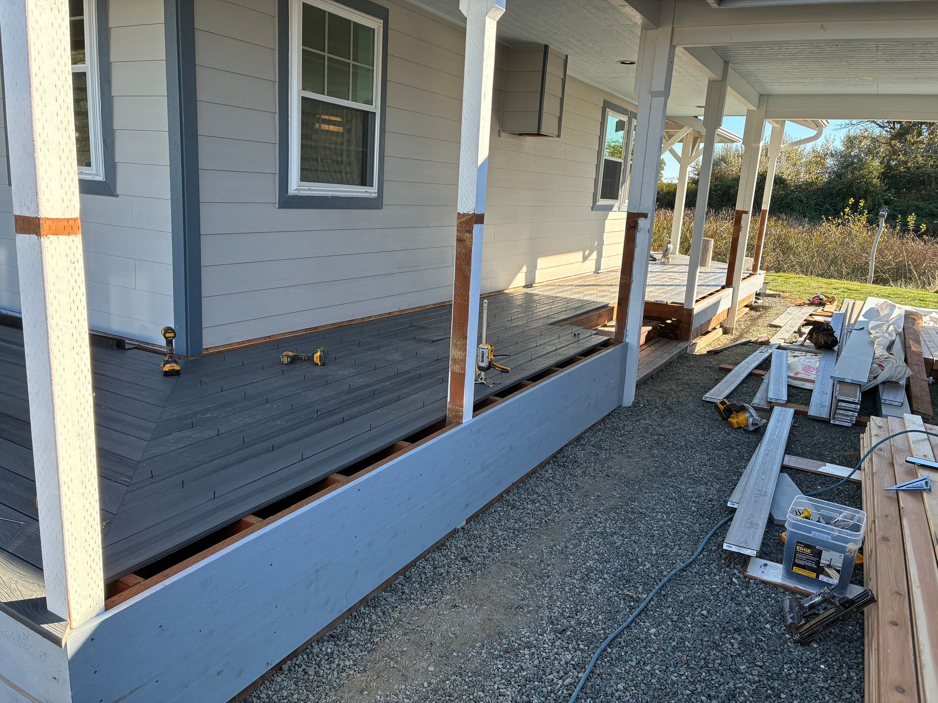 Partially built porch with white railings, gray siding, and construction materials along a gravel path