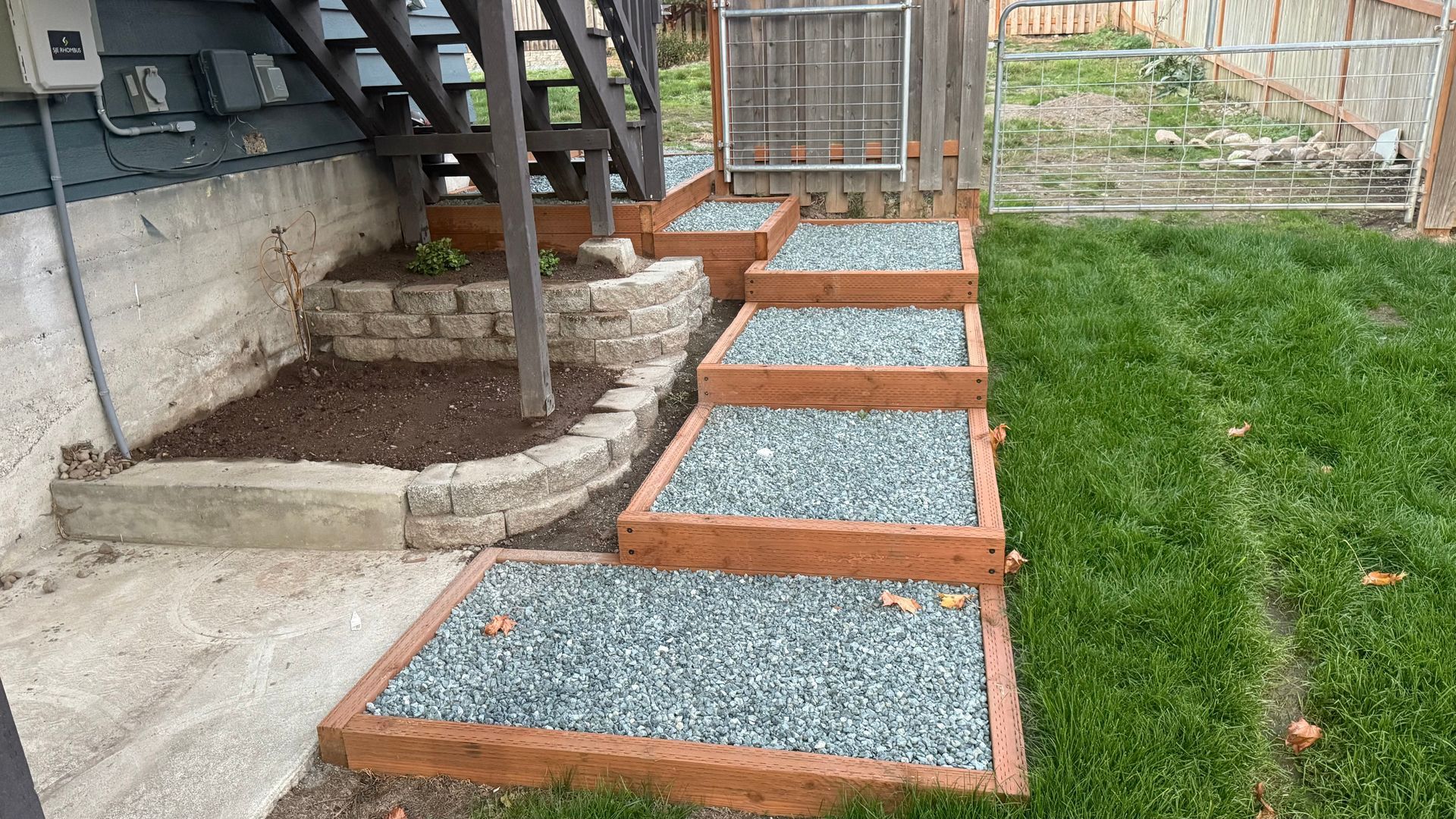 Gravel walkway with wooden edging leading beside a house to a fenced backyard and green lawn