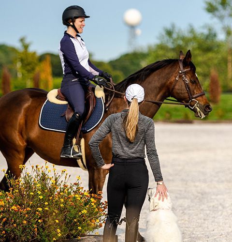 Woman on horseback in riding gear at an outdoor training area, with another person standing beside the horse