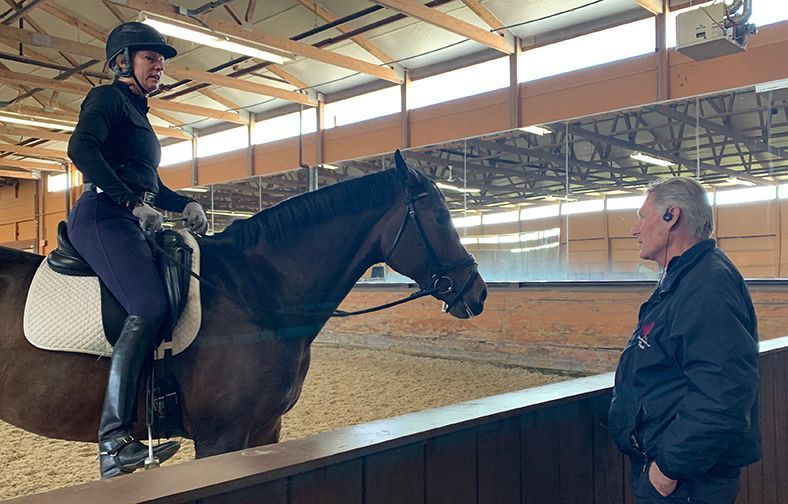 Rider on a dark horse indoors, speaking with an instructor beside the arena wall.