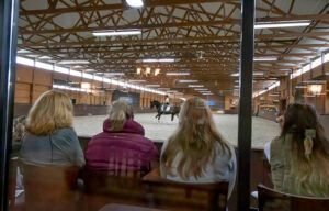 People seated on a bench watching an indoor skating rink with wooden beams and bright lights.