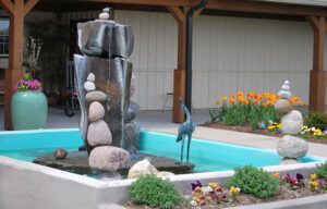Stone fountain with stacked rocks and flowing water beside a turquoise pool and flowers in a garden courtyard