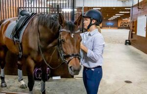 Person in a riding helmet standing beside a saddled horse in an indoor stable arena