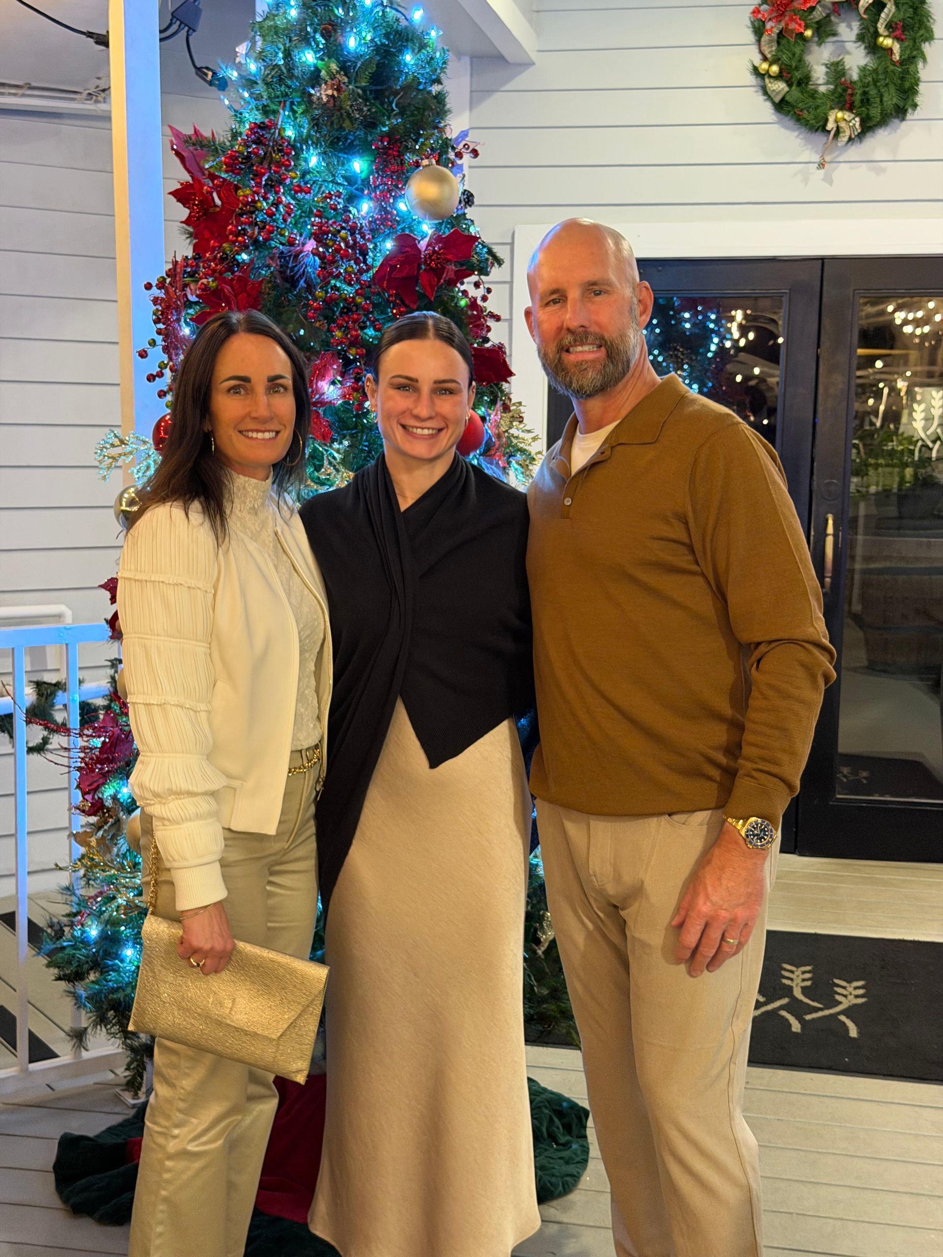 Three people smiling indoors beside a decorated Christmas tree and wreath.