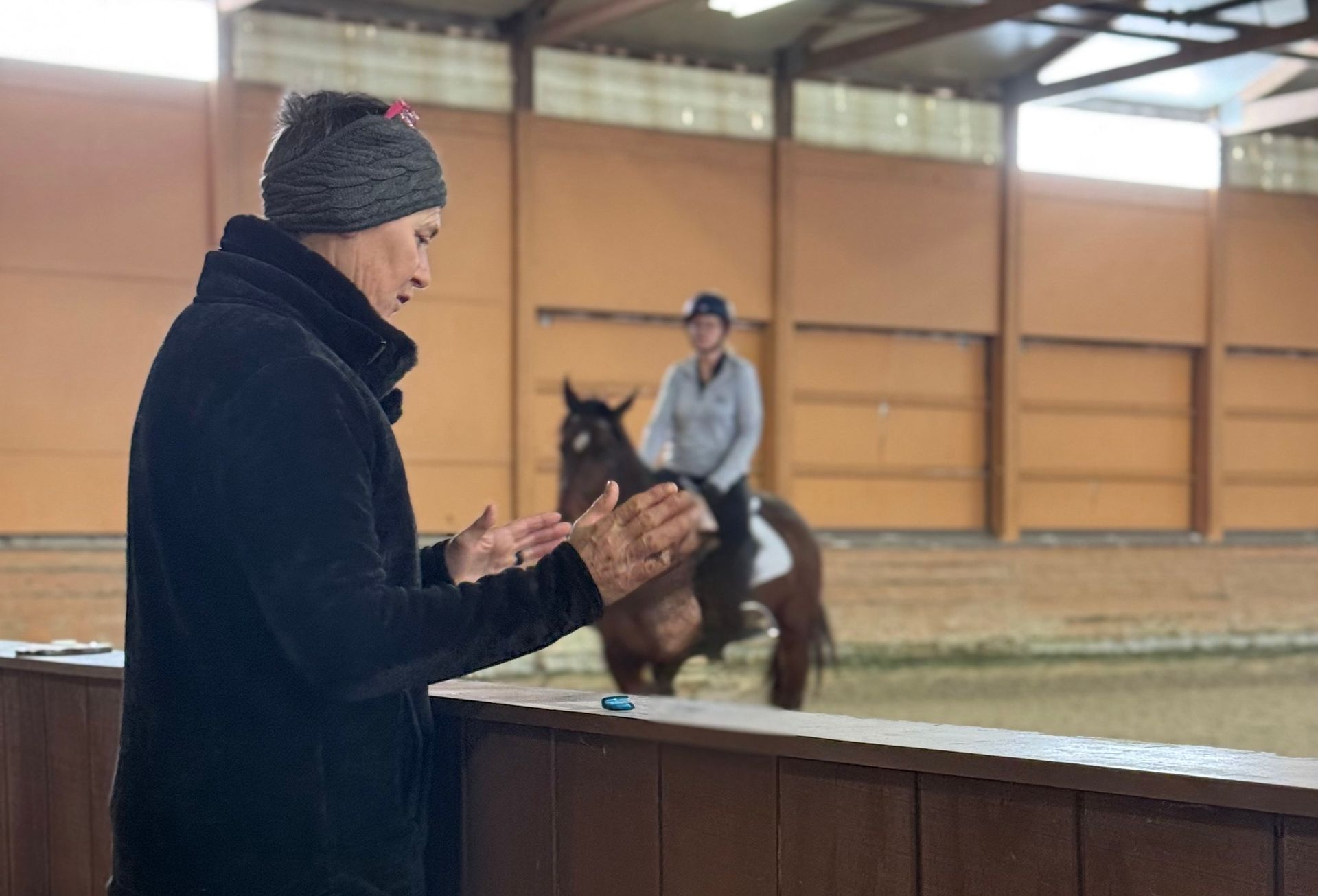 Person in winter clothes claps while watching a rider on a horse in an indoor arena