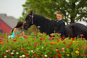 Man in suit on black horse beside woman in teal dress among red flowers outdoors