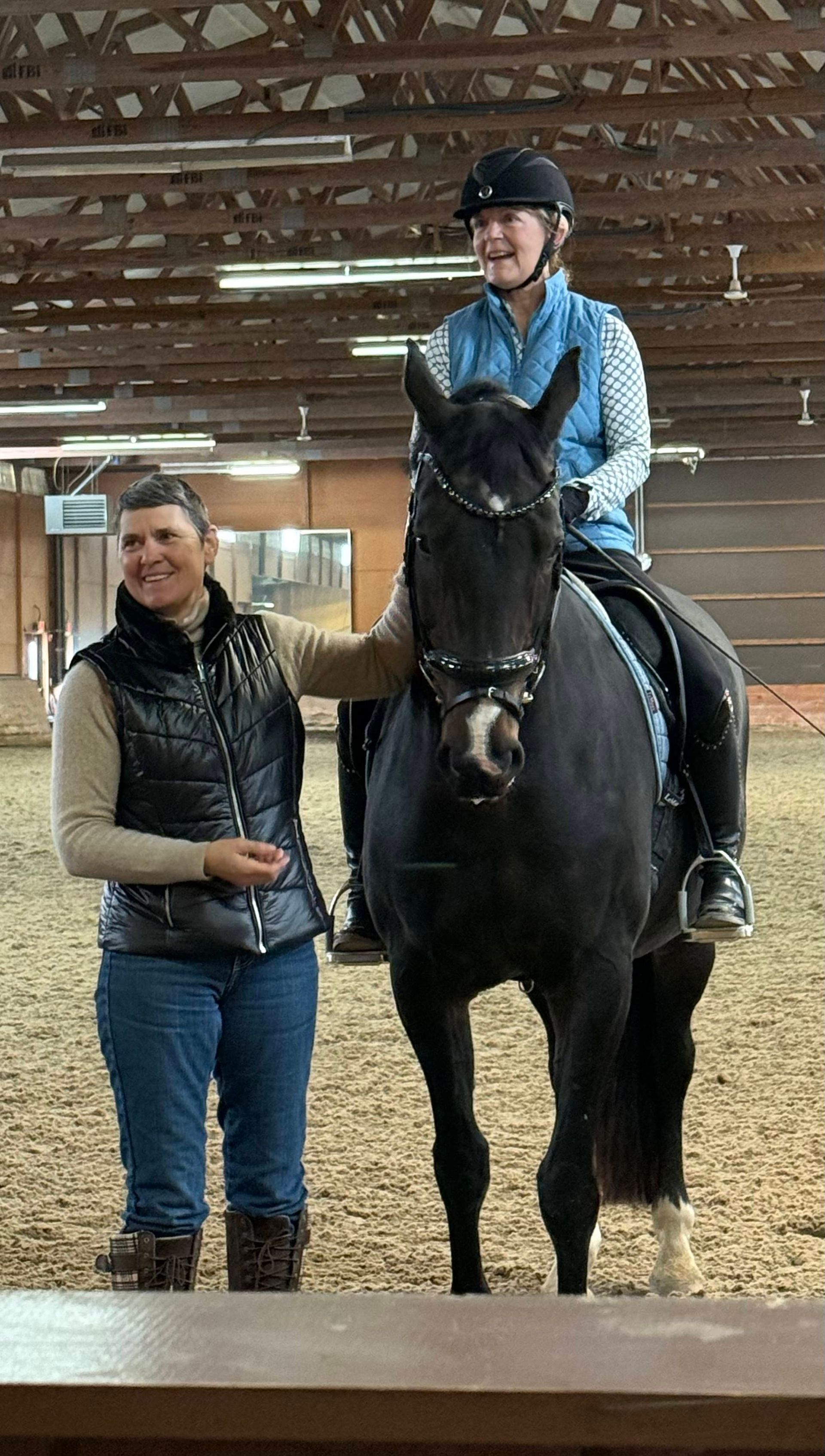 Person on a black horse with a handler in an indoor stable, both facing the camera.