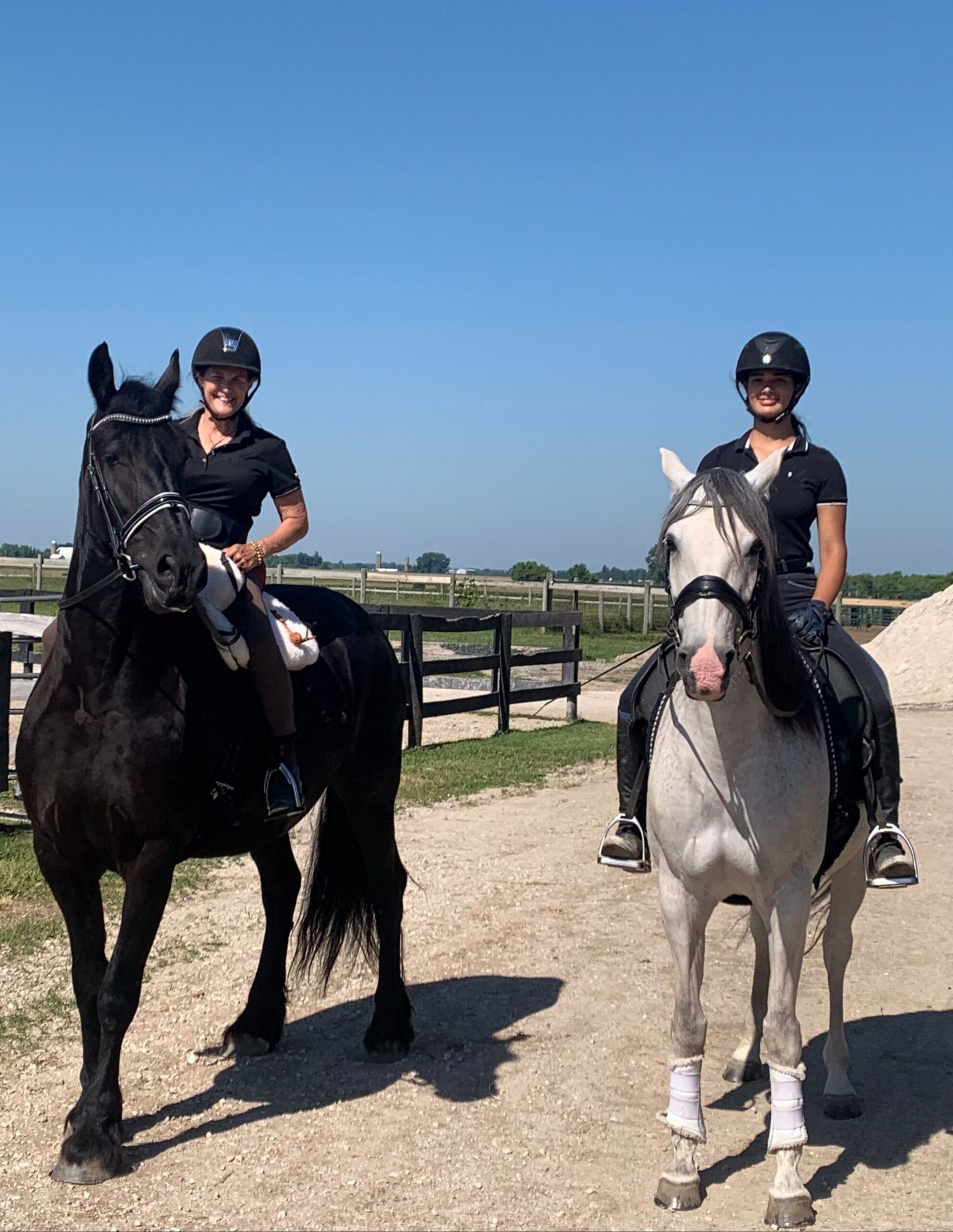 Two riders on horses outdoors under a clear blue sky, one black horse and one white horse.