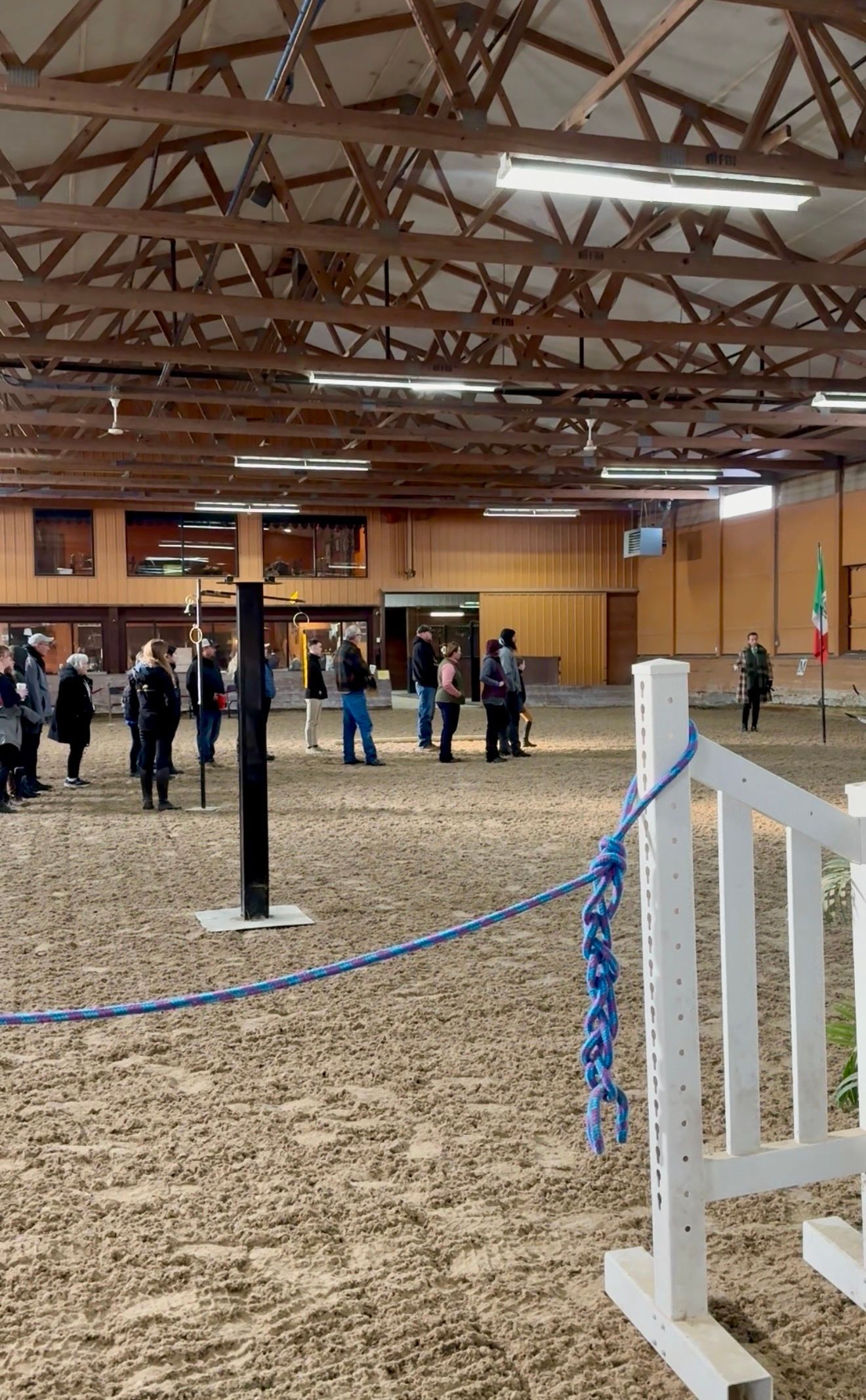 Indoor arena with a dirt floor, people gathered near a far wall, and white fence posts in the foreground