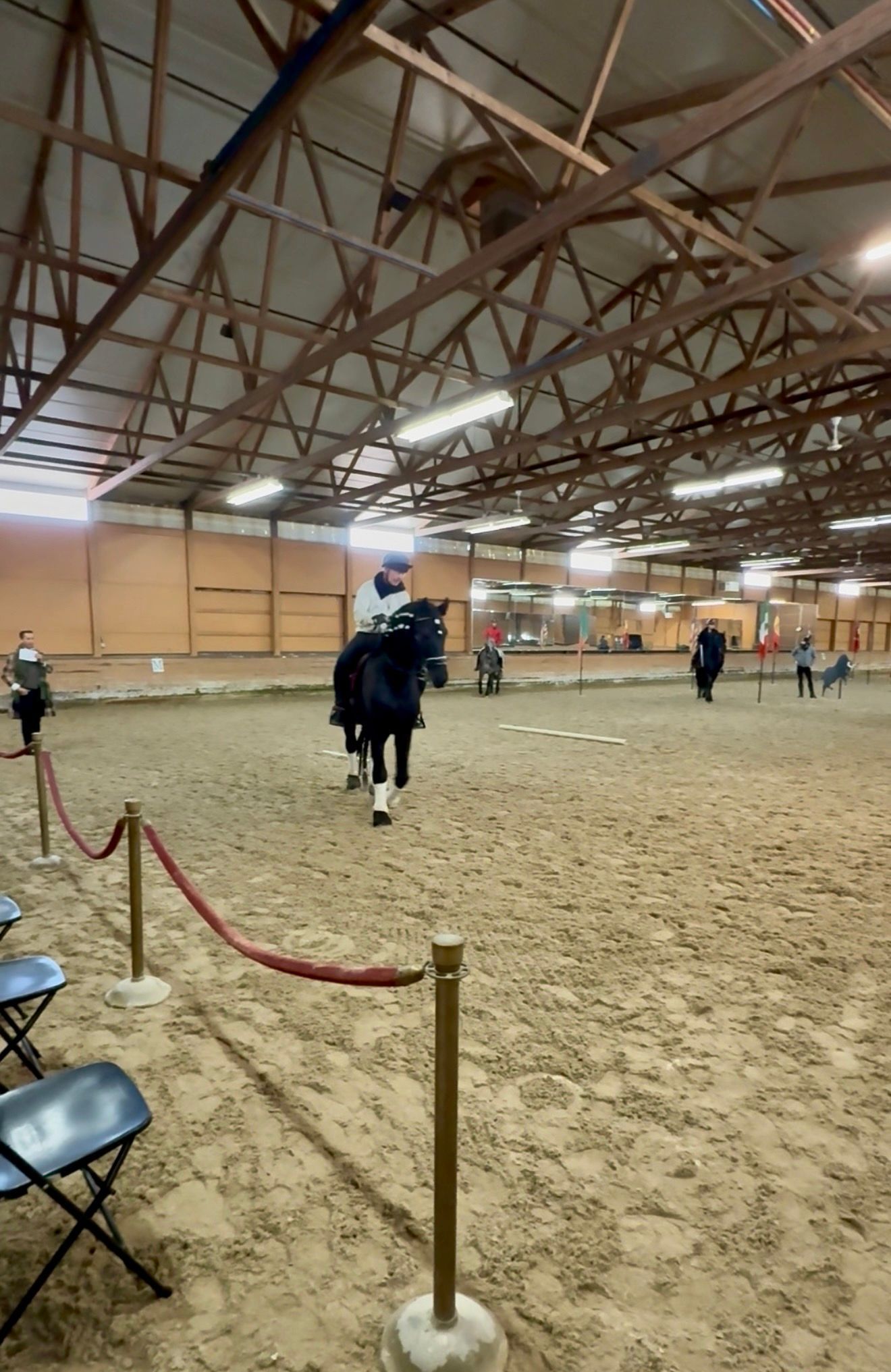 Indoor horse arena with a rider on a dark horse, red rope barrier, and spectators in the background