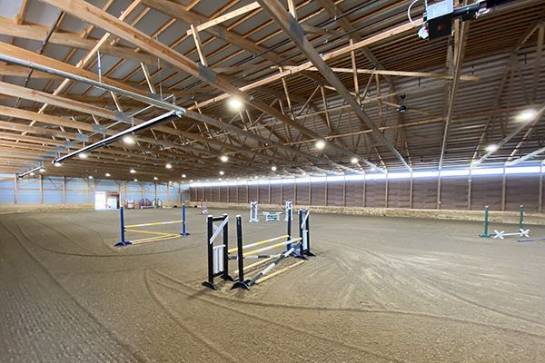 Empty indoor horse arena with jumps and sandy floor under wooden beams