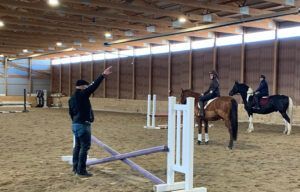 Indoor horse riding arena with riders and trainer over a low jump obstacle