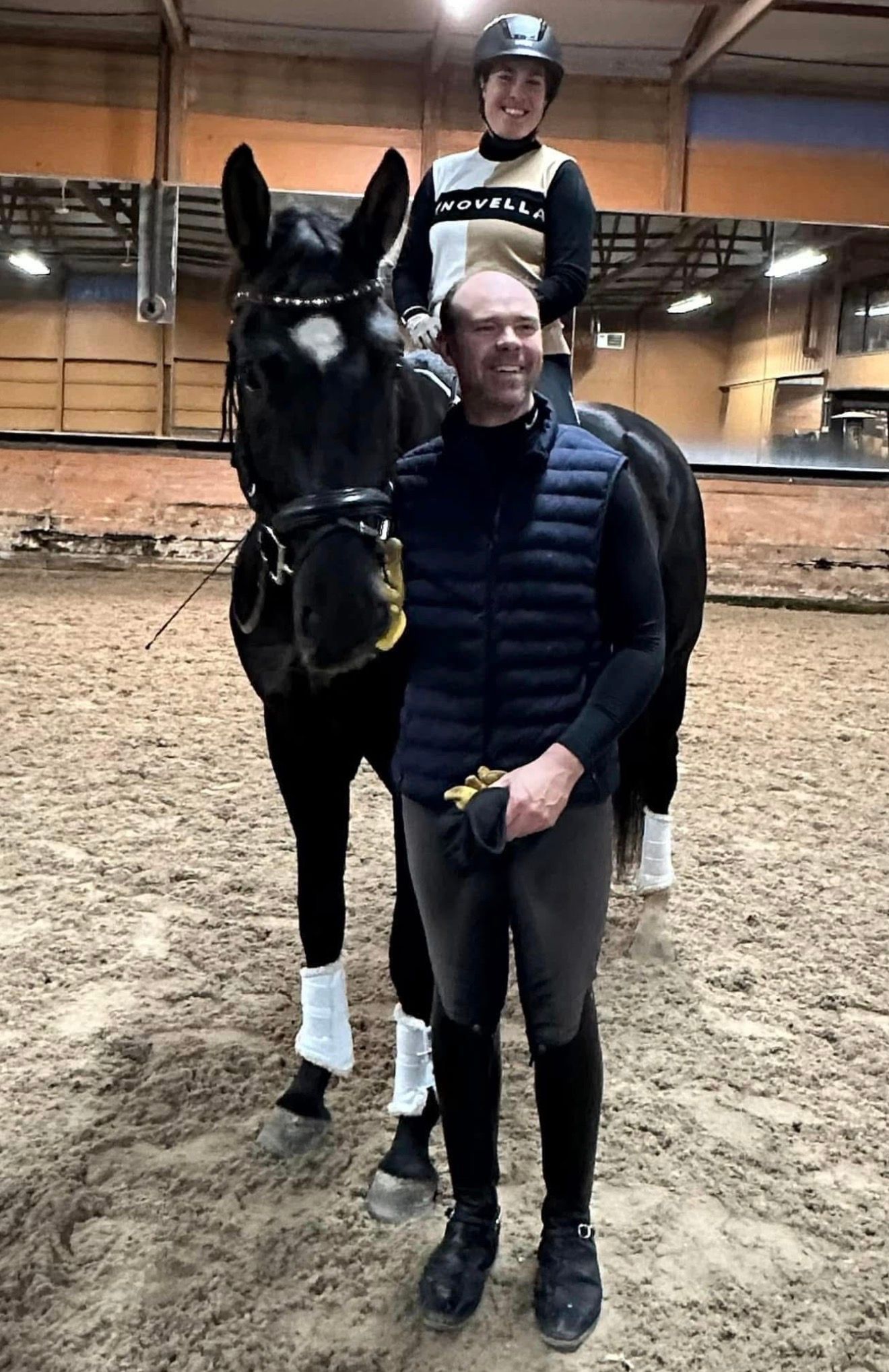 Person standing with a black horse indoors while a rider sits on its back