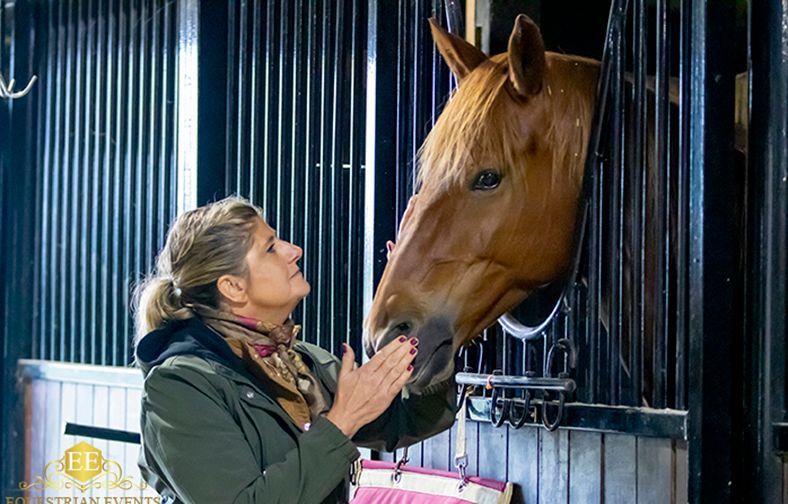 Woman gently touching a brown horse’s muzzle in a stable.