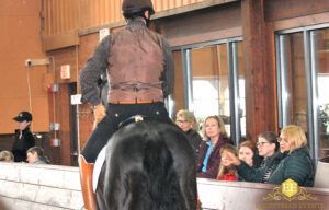 Mounted rider in brown vest on a black horse indoors beside spectators in a viewing area