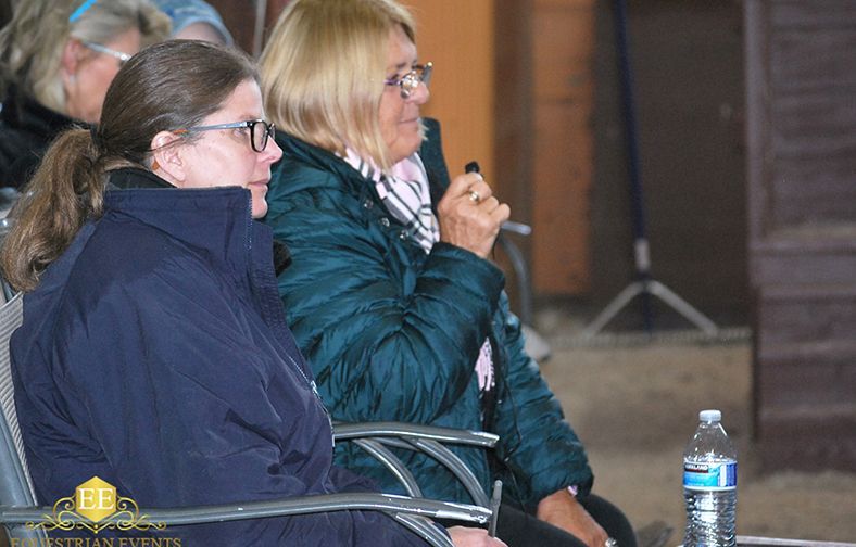 Two women in winter jackets sit outdoors, one holding a microphone beside a water bottle.