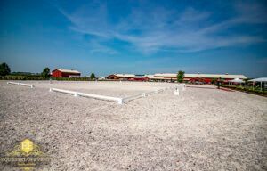 Wide gravel equestrian arena with white rails under a blue sky, red barns in the distance