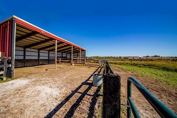 Open-sided red barn beside a fenced dirt path under a bright blue sky