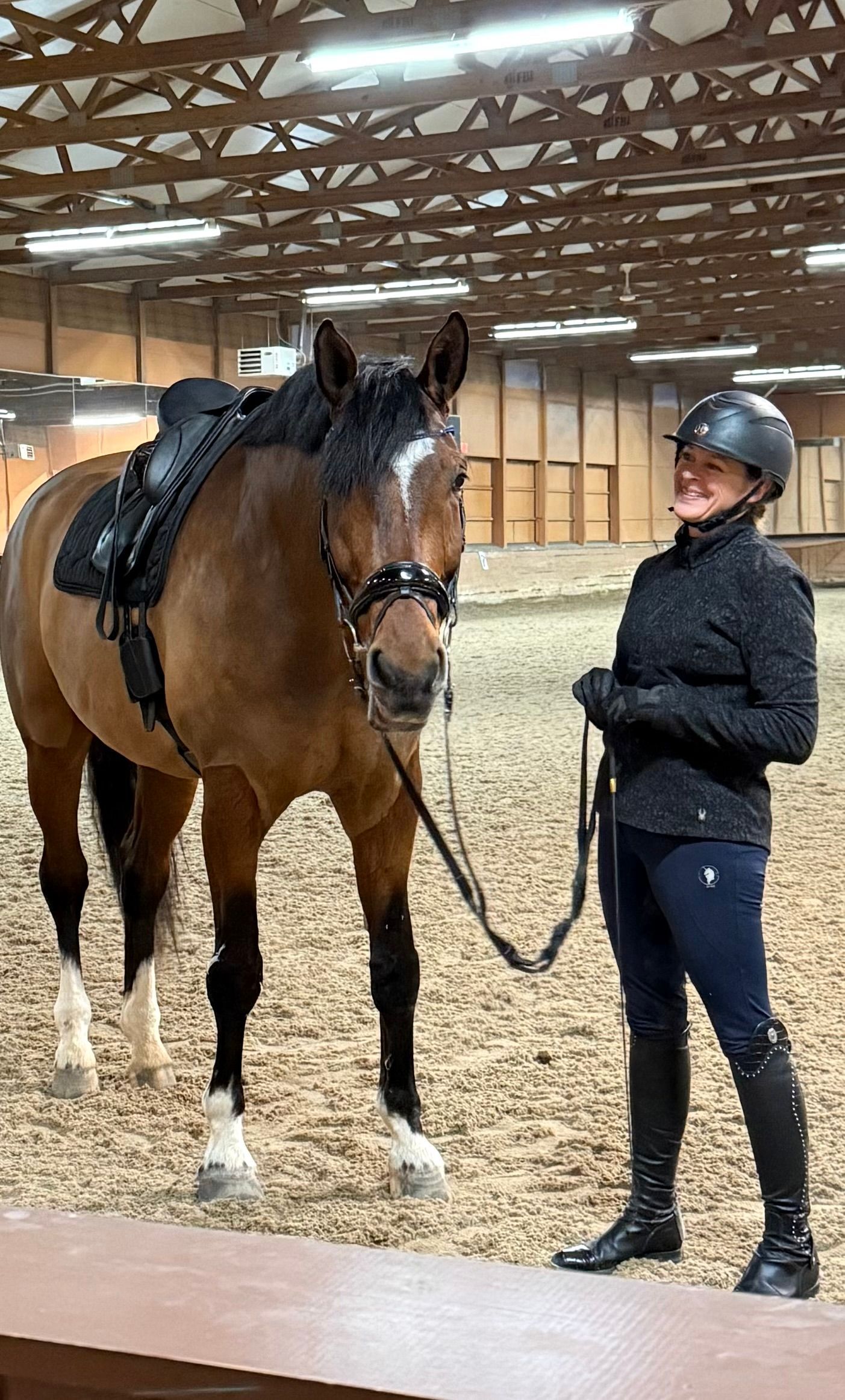 Person in riding helmet standing beside saddled brown horse in an indoor arena