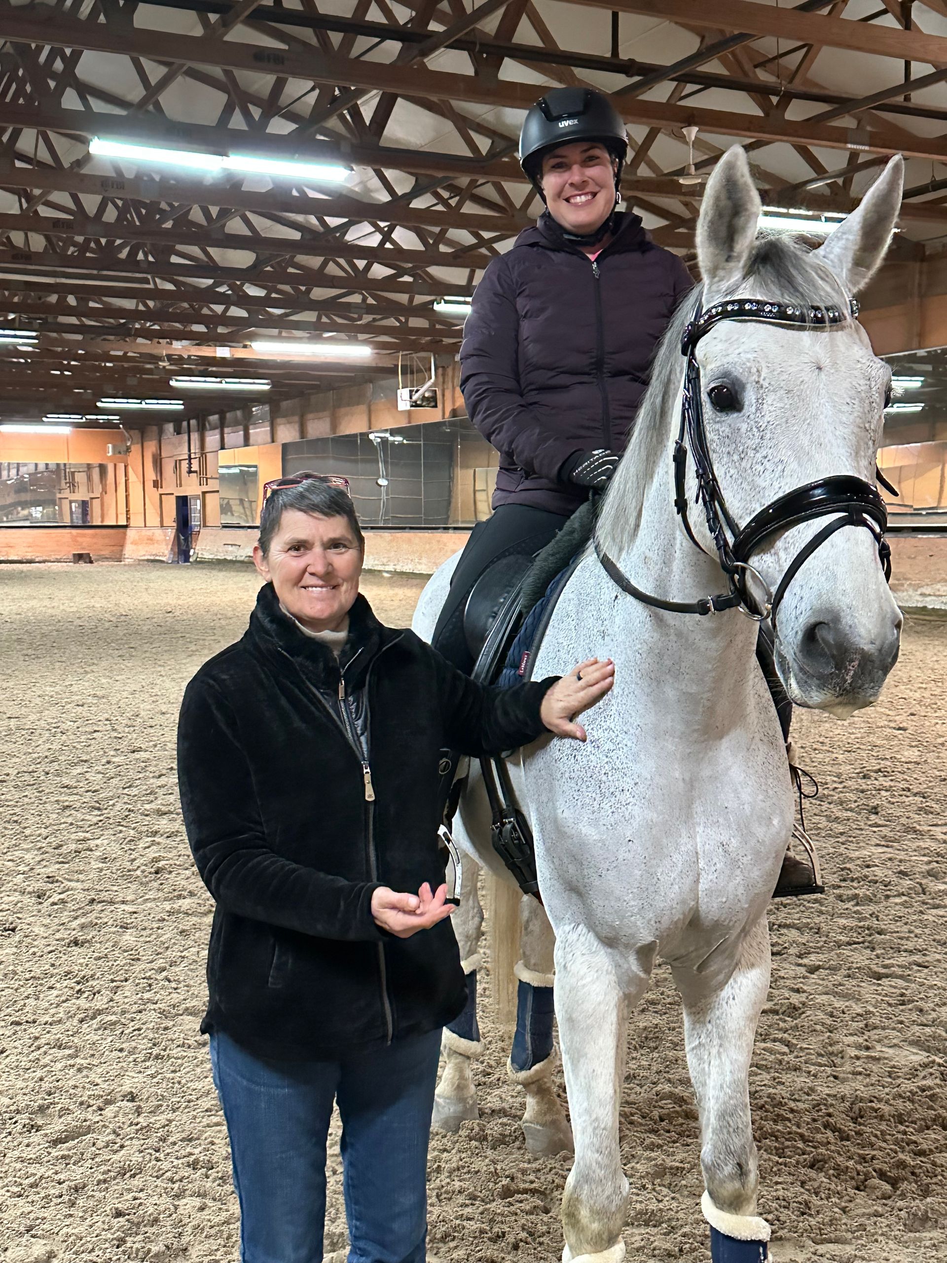 Two people with a gray horse in an indoor riding arena, one mounted and one standing beside it.