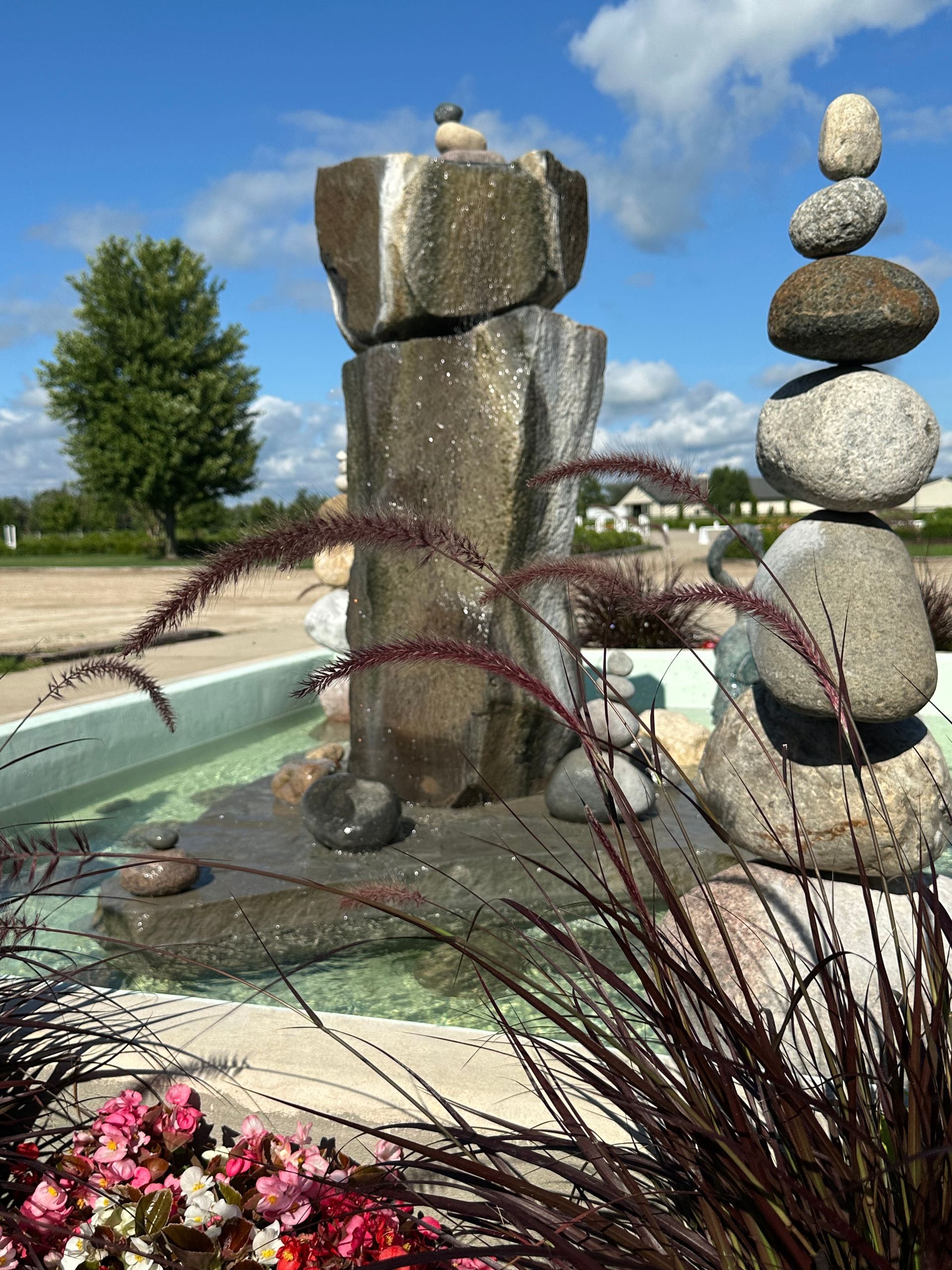 Outdoor fountain with stacked stone sculptures, flowers, and ornamental grasses under a blue sky