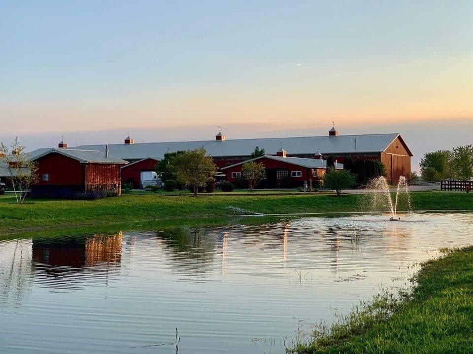 Red barns by a pond at sunset, with a fountain spraying and reflections in the water