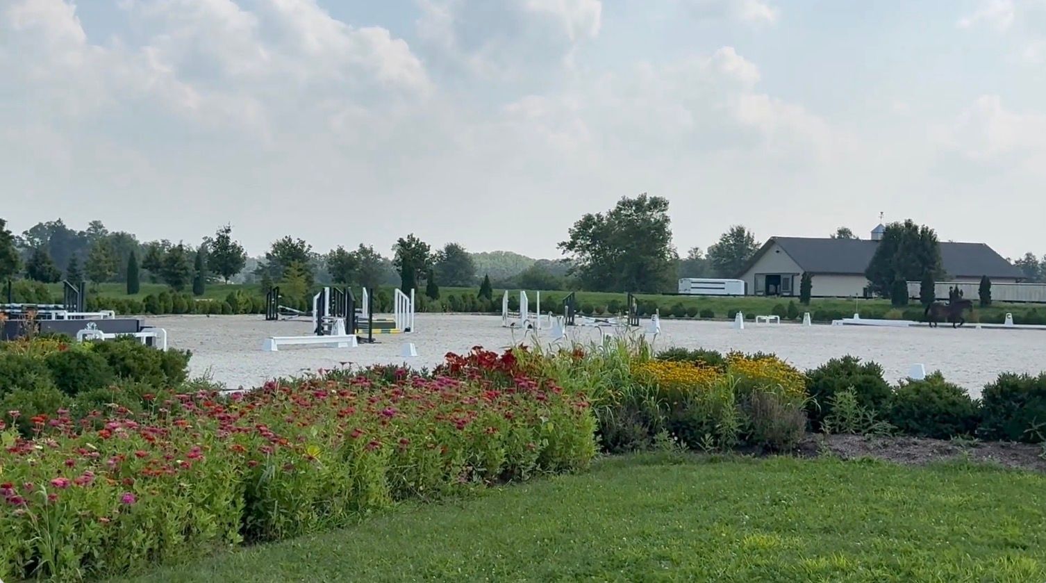 Flower beds beside a grassy park path, with a fountain and houses in the distance under a cloudy sky