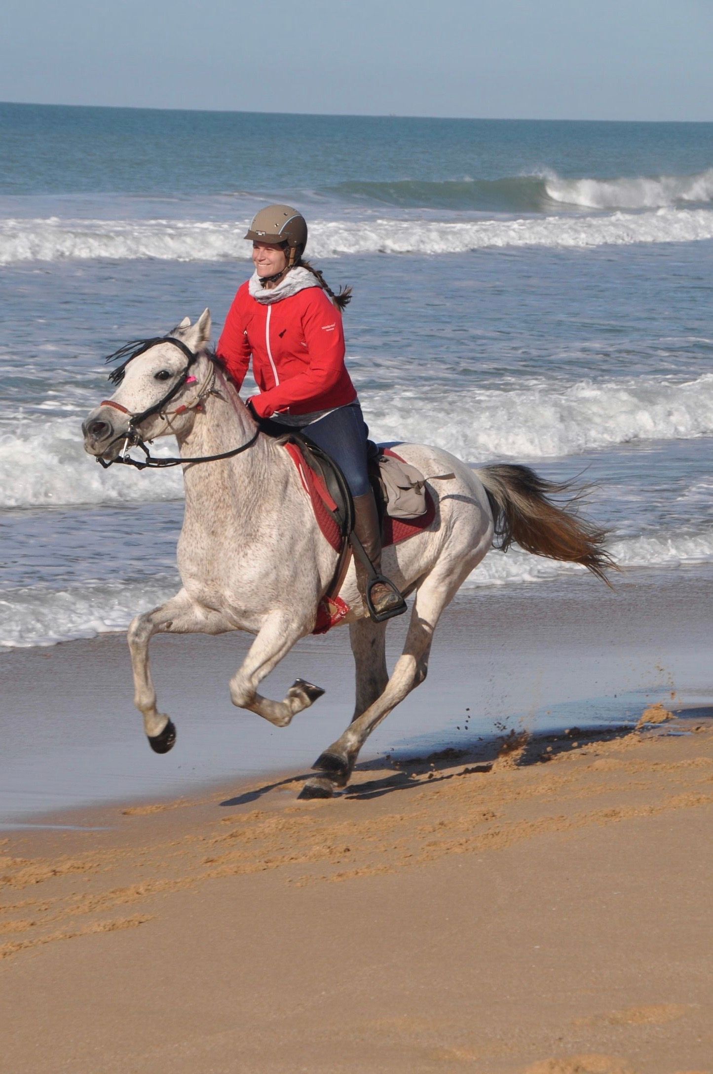 Person in a red jacket riding a white horse along a beach with waves in the background