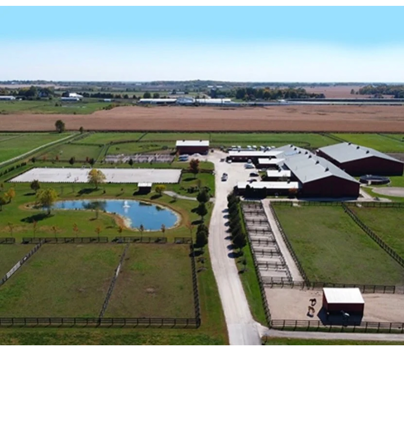Aerial view of a rural farm with barns, fenced fields, a driveway, and a small pond