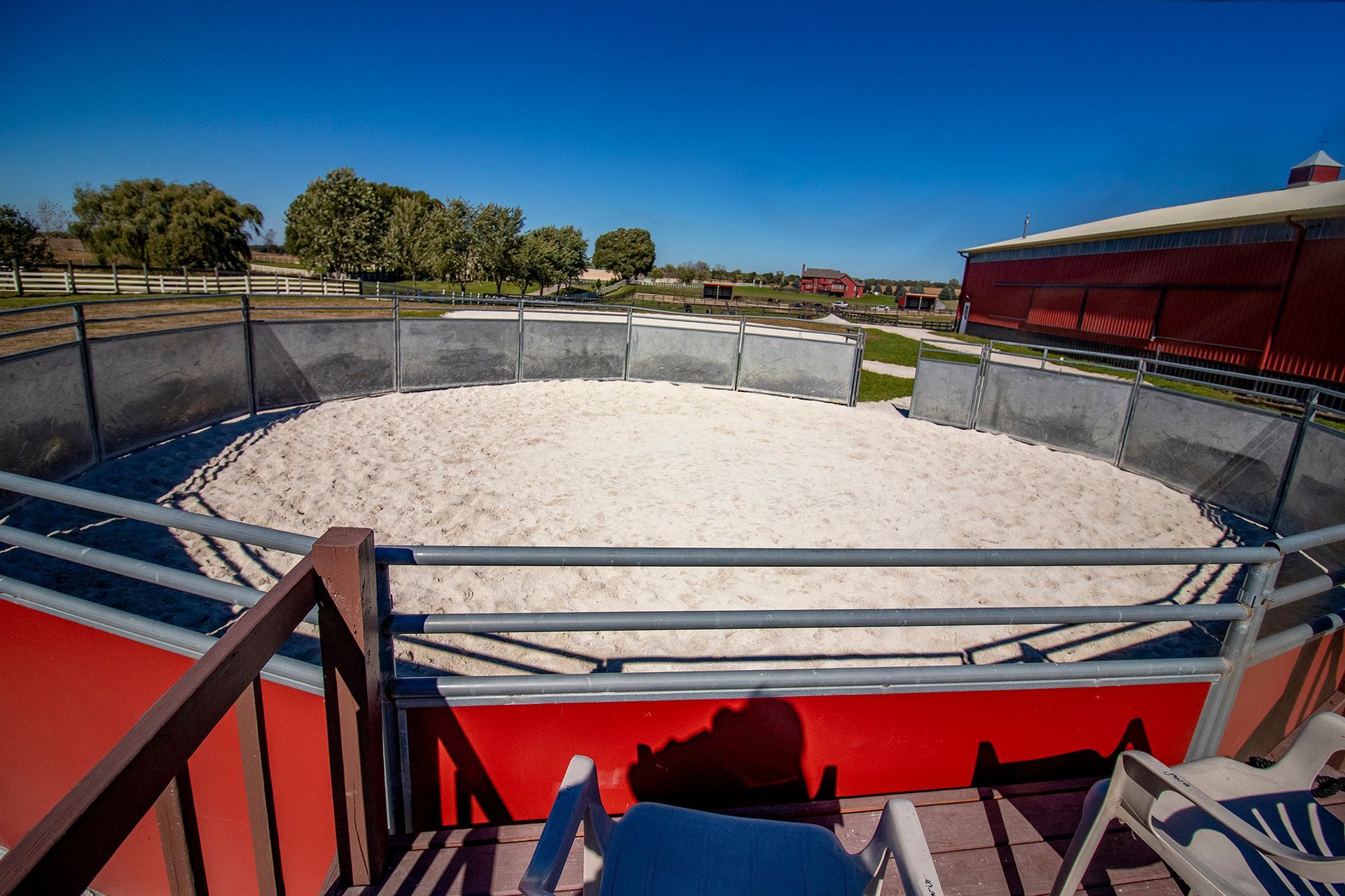 Outdoor rodeo arena with a white dirt ring, metal fencing, and red spectator seating under a clear blue sky