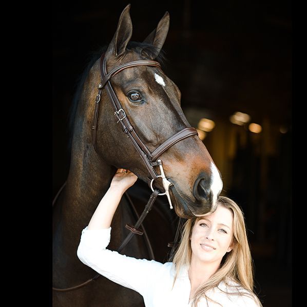 Woman in white stands beside a dark horse in a stable doorway, holding its bridle.