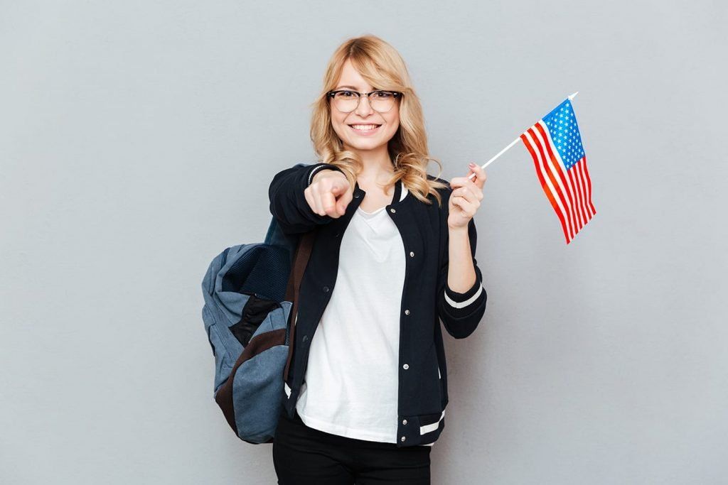 Smiling person holding a small American flag and pointing, with a backpack against a gray background