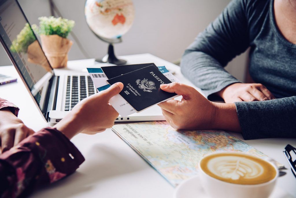 Two people exchange a passport over a laptop beside a coffee cup and map at a travel planning table