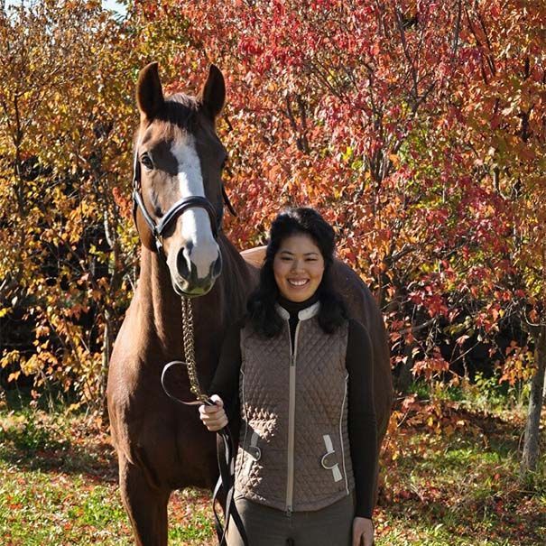 Woman standing beside a brown horse in an autumn park, with colorful fall foliage behind them.