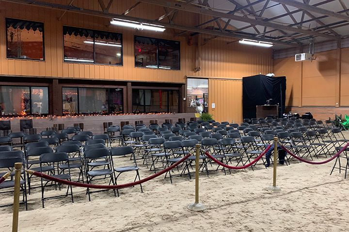 Empty indoor arena with rows of folded chairs on sand, rope barriers, and a wooden wall with windows.