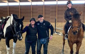 Two people standing between two horses in an indoor arena, with one rider on each horse.