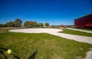 Grassy field with a paved path, trees in the distance, and a red building under a blue sky