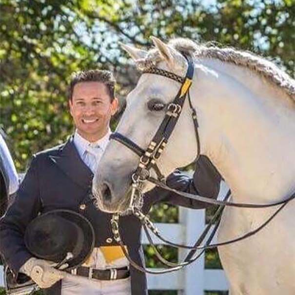 Man in a dark suit standing beside a white horse with a black bridle outdoors